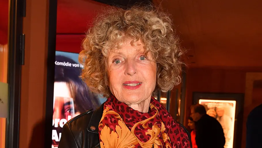 Kathrin Ackermann Eine Frau mit lockigem Haar trägt eine schwarze Lederjacke und einen rot-orange gemusterten Schal. Hinter ihr hängen Plakate.