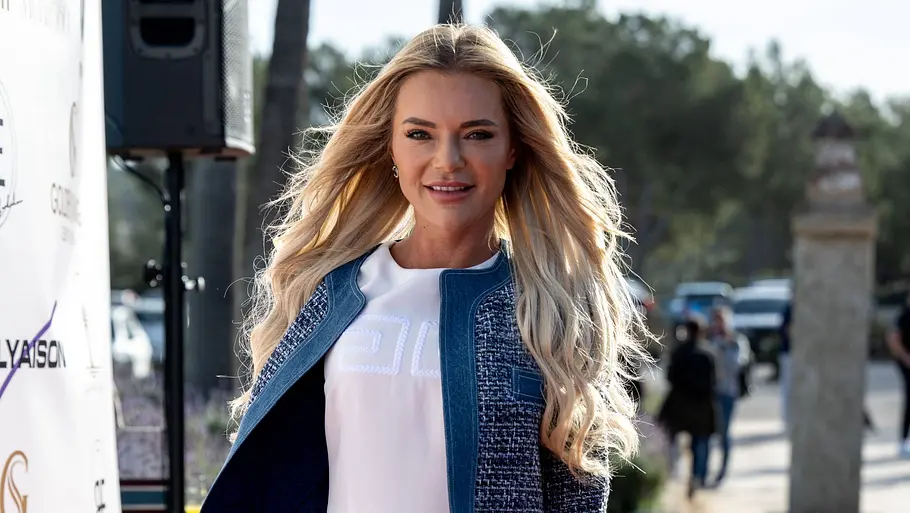 Eine blonde Frau läuft lächelnd über einen roten Teppich im Freien. Sie trägt ein weißes Kleid, eine kurze Jacke und hohe weiße Stiefel, im Hintergrund sind Event-Logos und Zuschauer zu sehen.
