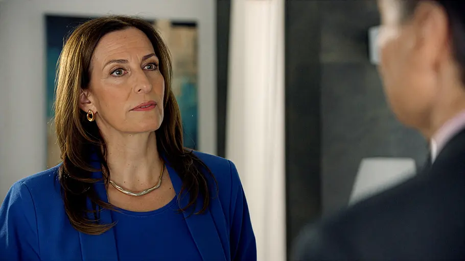 Katrin trägt einen blauen Blazer und goldenen Schmuck. Sie blickt Gerner an, der rechts unscharf im Vordergrund steht.