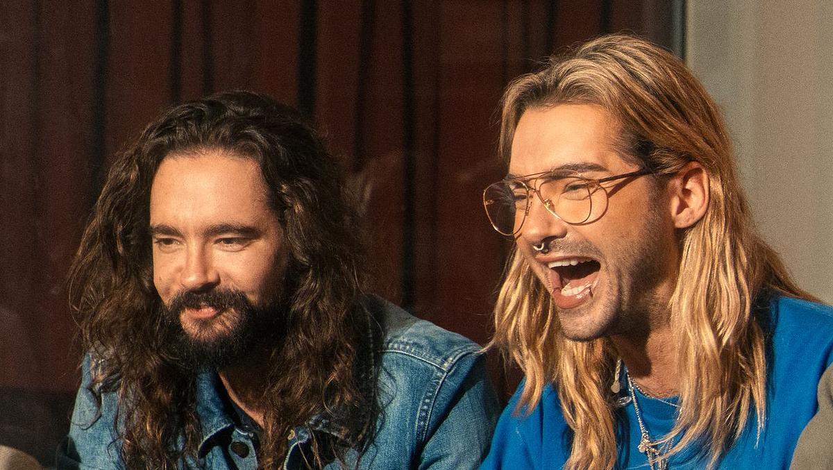 „Kaulitz & Kaulitz“, Staffel 3 Tom Kaulitz sitzt in Jeans-Outfit auf dem Sofa, neben ihm sein mit offenem Mund lachender Bruder Bill Kaulitz mit Brille und blauem Pulli