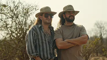 Bill und Tom Kaulitz stehen gemeinsam in einer trockenen Savannenlandschaft. Beide tragen helle Outdoor-Hüte, während Bill mit Sonnenbrille und gestreiftem Hemd zu sehen ist und Tom ein beiges T-Shirt trägt. Im Hintergrund sind kahle Bäume und Buschlandschaft zu erkennen. - Foto: Netflix