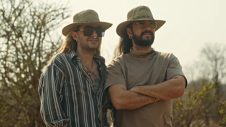Bill und Tom Kaulitz Bill und Tom Kaulitz stehen gemeinsam in einer trockenen Savannenlandschaft. Beide tragen helle Outdoor-Hüte, während Bill mit Sonnenbrille und gestreiftem Hemd zu sehen ist und Tom ein beiges T-Shirt trägt. Im Hintergrund sind kahle Bäume und Buschlandschaft zu erkennen.