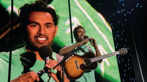 Jack Malik steht mit Gitarre und Mikrofon auf der Bühne, sein Gesicht ist hinter ihm auch auf einer großen Leinwand zu sehen. - Foto: TMDB