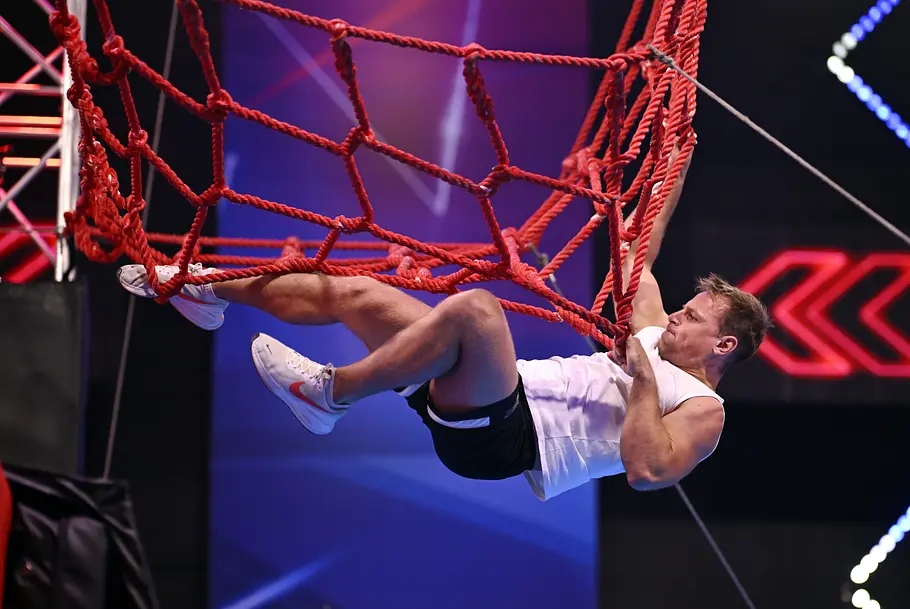 Kevin Frey ist Teilnehmer bei Ninja Warrior Germany 2026 Kevin Frey hängt in weißem Shirt und schwarzer Shorts an einem roten Kletternetz im beleuchteten Hindernisparcours.