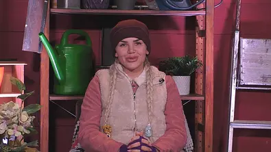 Kim Virginia sitzt mit Mütze, zwei Zöpfen und heller Weste vor einem Holzregal mit grüner Gießkanne und Topfpflanze. - Foto: Joyn Kim Virginia sitzt mit Mütze, zwei Zöpfen und heller Weste vor einem Holzregal mit grüner Gießkanne und Topfpflanze. - Foto: Joyn