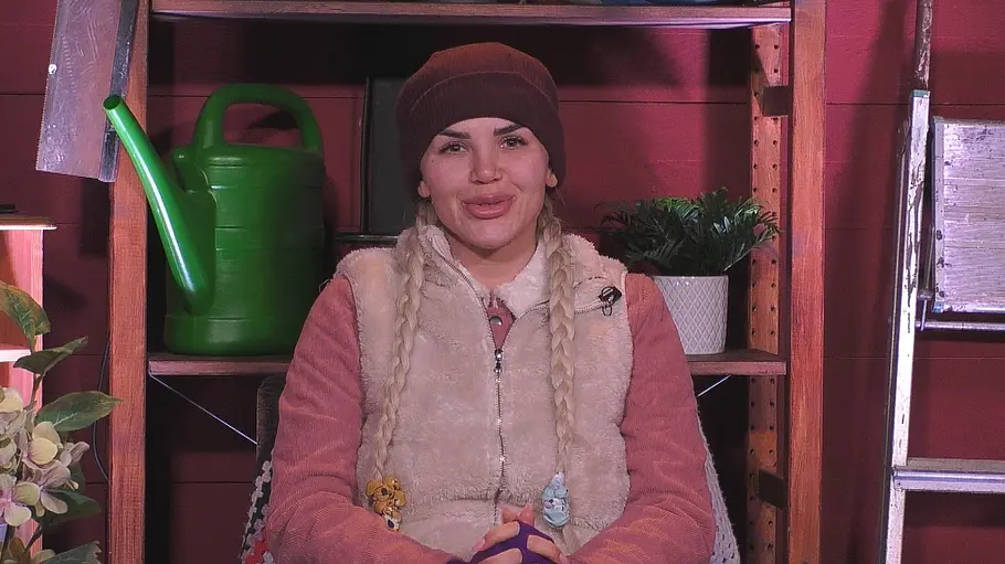Kim Virginia Kim Virginia sitzt mit Mütze, zwei Zöpfen und heller Weste vor einem Holzregal mit grüner Gießkanne und Topfpflanze.