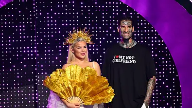 Kim Virginia mit goldener Krone und großem Goldfächer steht neben dem tätowierten Nikola vor einer violetten Lichtwand. - Foto: Joyn