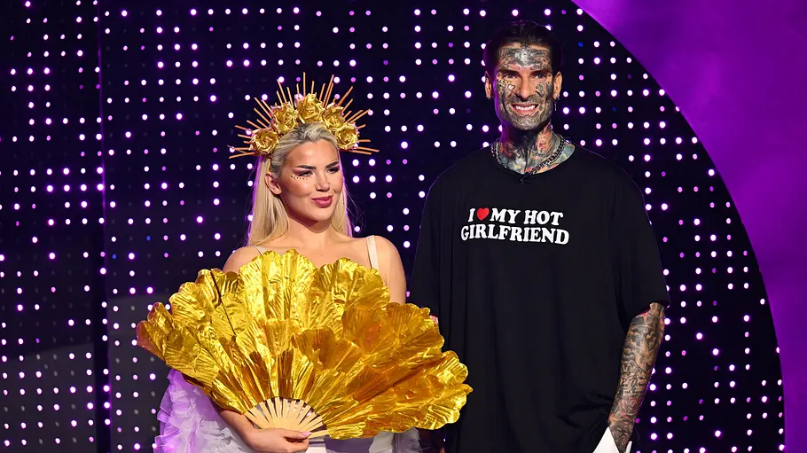 Kim Virginia und Nikola Kim Virginia mit goldener Krone und großem Goldfächer steht neben dem tätowierten Nikola vor einer violetten Lichtwand.