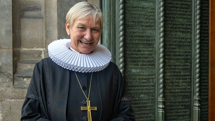 Kirsten Fehrs Kirsten Fehrs lächelt im schwarzen Talar mit weißer Halskrause und goldenem Kreuz vor einer beschrifteten Metalltür.