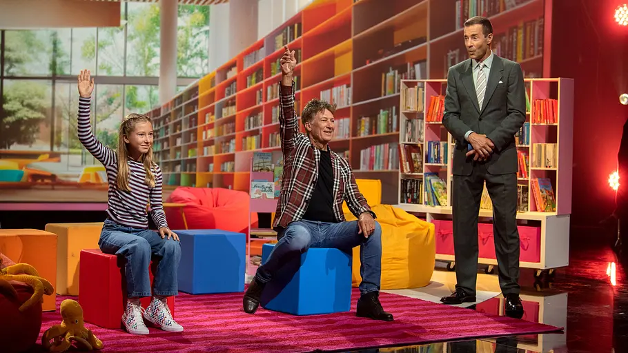 Tobias Moretti bei „Klein gegen Groß“ Tobias Moretti sitzt mit seiner jungen Herausforderin Marlies vor einer Bücherkulisse bei „Klein gegen Groß“, während Kai Pflaume danebensteht.