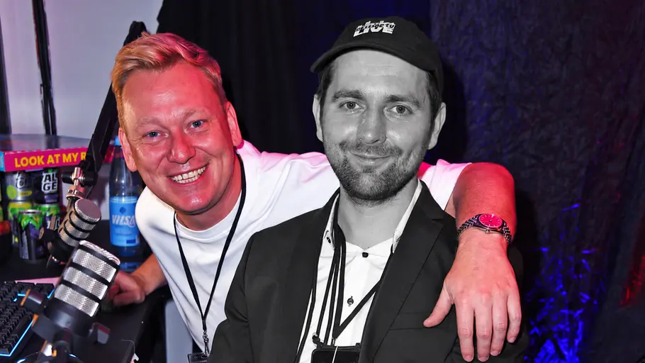 Letzte Adam-Wolke-Streams: Kopfschmerzen Knossi legt den Arm um Adam Wolke, der schwarz-weiß dargestellt ist. Beide lächeln neben Mikrofonen in die Kamera.