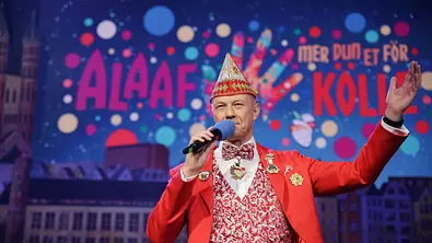 Moderator im roten Karnevalskostüm mit Mikrofon auf der Bühne, im Hintergrund bunte „Alaaf“-Kulisse beim Kölner Karneval. - Foto: WDR/Dirk Borm