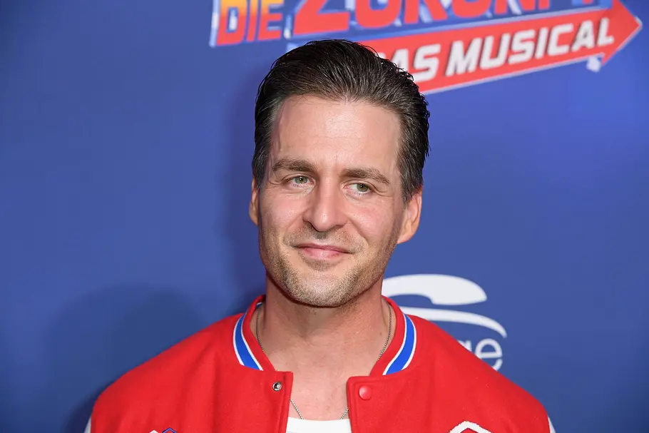 Alexander Klaws trägt eine rote Collegejacke und lächelt vor einer blauen Wand mit einem Musical-Schriftzug.