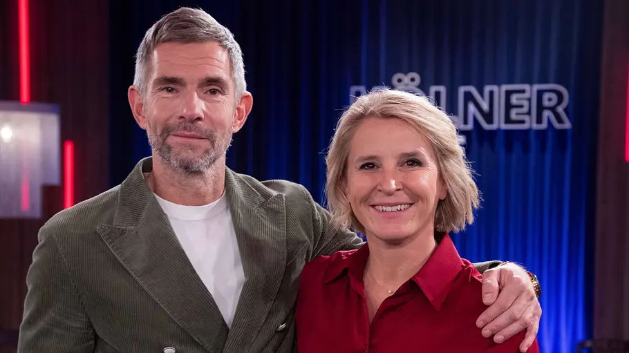 Susan Link und Micky Beisenherz Micky Beisenherz im grünen Sakko legt den Arm um Susan Link in roter Bluse. Beide stehen vor einem blauen Vorhang.