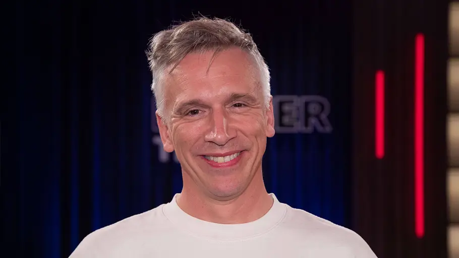 Bastian Campmann Bastian Campmann lächelt in einem weißen T-Shirt vor einem dunklen Hintergrund mit blauen und roten Lichtstreifen.