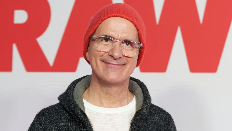 Christoph Maria Herbst Mann mit roter Strickmütze und transparenter Brille lächelt vor einer weißen Fotowand mit großen roten Schriftzügen. Er trägt ein weißes T-Shirt und eine dunkelgraue Strickjacke mit hellen und roten Akzenten.