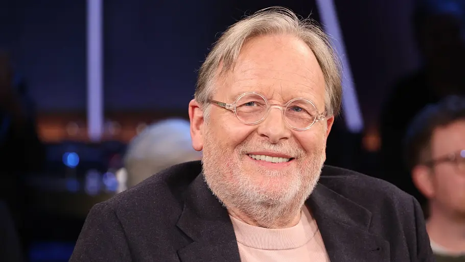 Ein älterer Mann mit grauem Haar, Bart und runder Brille sitzt in einem TV-Studio und lächelt freundlich in die Kamera, während im Hintergrund unscharf weitere Gäste zu sehen sind.