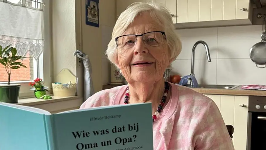 Elfriede Heitkamp Ältere Frau mit Brille sitzt in ihrer Küche und hält ein Buch mit dem Titel „Wie was dat bij Oma un Opa?“ in die Kamera. Im Hintergrund sind ein Fenster, Pflanzen und Kücheneinrichtung zu sehen.
