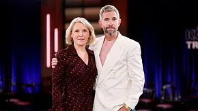 Zwei Personen stehen eng beieinander in einem TV-Studio und blicken in die Kamera. Die Frau trägt ein dunkel gemustertes Kleid, der Mann ein helles Sakko mit Jeans, während im Hintergrund die Studiokulisse mit blauer Beleuchtung zu sehen ist. - Foto: IMAGO / Panama Pictures
