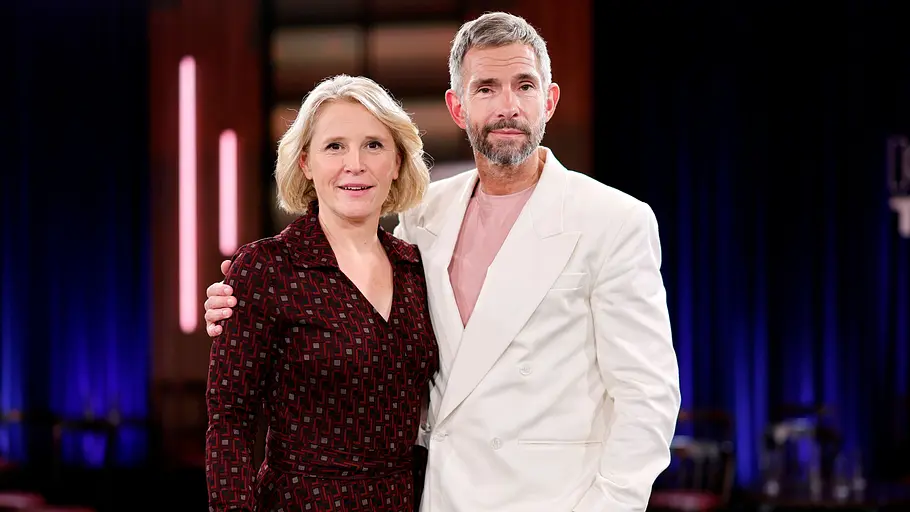 Kölner Treff Zwei Personen stehen eng beieinander in einem TV-Studio und blicken in die Kamera. Die Frau trägt ein dunkel gemustertes Kleid, der Mann ein helles Sakko mit Jeans, während im Hintergrund die Studiokulisse mit blauer Beleuchtung zu sehen ist.