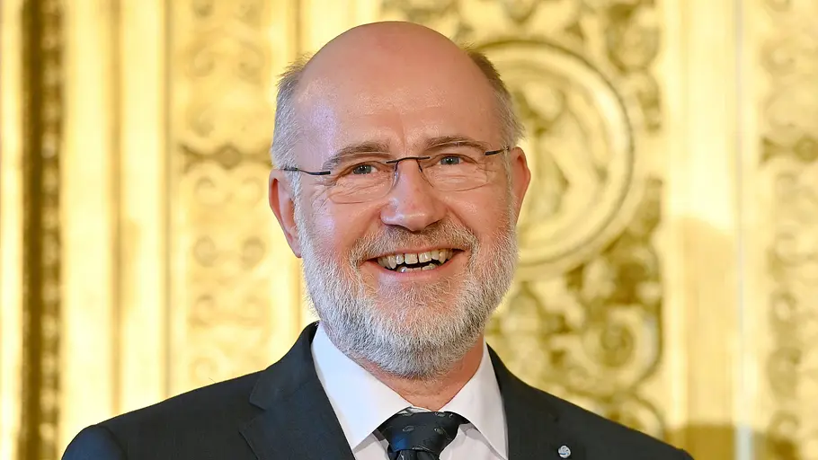 Harald Lesch Porträt eines lächelnden Mannes mit Brille, grauem Bart und dunklem Anzug vor einem dekorativen, goldfarbenen Hintergrund.