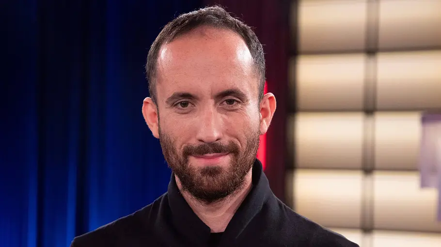 Igor Levit Igor Levit trägt eine schwarze Jacke und lächelt in die Kamera. Hinter ihm sind blaue Vorhänge und helle Leuchtflächen.