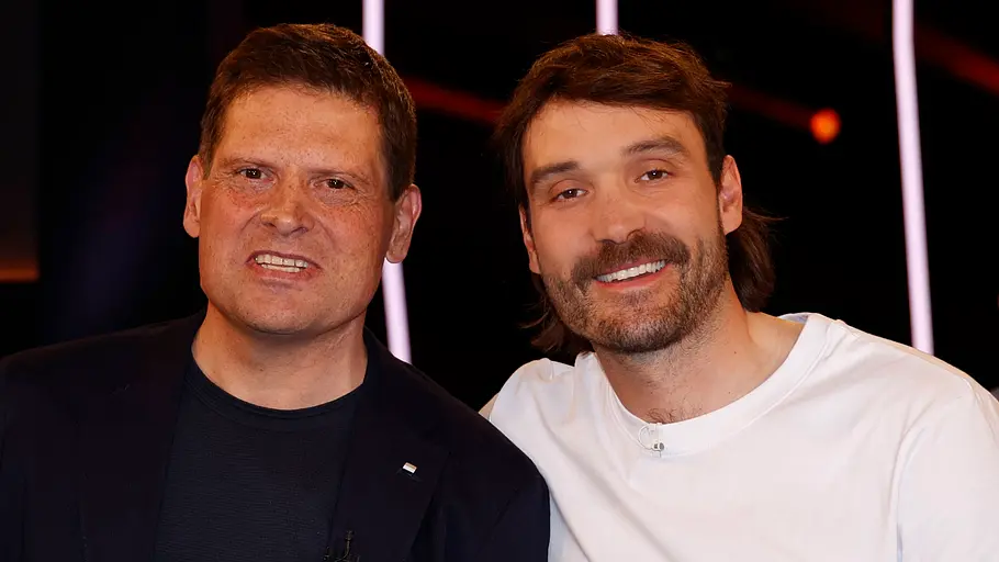 Zwei Männer stehen eng nebeneinander in einem TV-Studio und lächeln in die Kamera, im Hintergrund sind dunkle Bühnenlichter und eine Showkulisse zu erkennen.