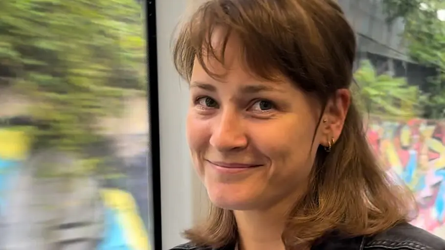 Jennifer Springer Jennifer Springer sitzt in dunkler Jeansjacke am Fenster einer Bahn und lächelt. Draußen sind Bäume und Graffiti zu sehen.