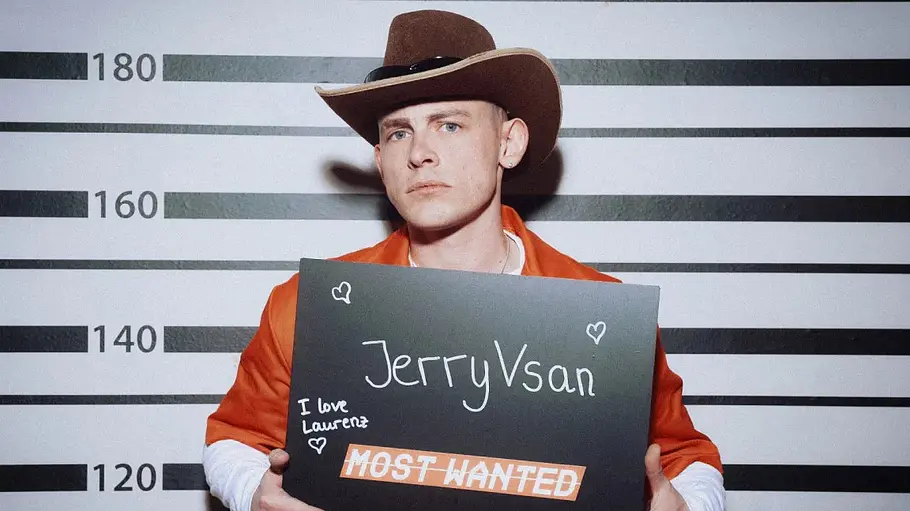 Ein Mann mit ernstem Blick trägt einen Cowboyhut und einen orangefarbenen Overall und steht vor einer Wand mit Höhenmarkierungen wie bei einem Polizeifoto. In den Händen hält er ein Schild mit der Aufschrift „JerryVsan“ und „Most Wanted“, verziert mit kleinen Herzzeichnungen.