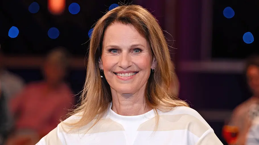 Eine Frau mit schulterlangem Haar lächelt in die Kamera und steht in einem TV-Studio, im Hintergrund sind unscharf Publikum und Lichter zu sehen.