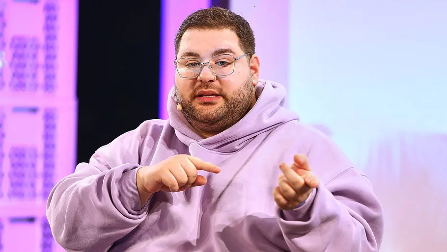 Leon xSkincare Mann mit Brille und hellem Kapuzenpullover spricht auf einer Bühne und gestikuliert mit den Händen, während er etwas erklärt. Im Hintergrund sind rosa und violett beleuchtete Elemente einer Studiokulisse zu sehen.