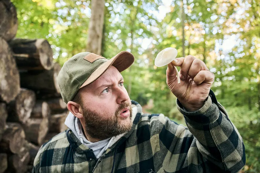 Martin Maschka trägt eine Kappe und eine karierte Jacke. Er hält im Wald einen hellen Pilz hoch und betrachtet ihn.