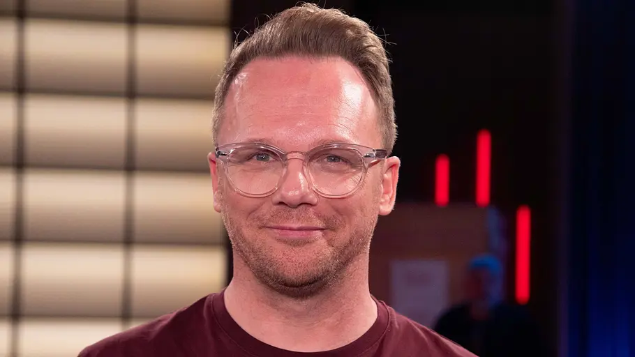 Ralf Schmitz Ralf Schmitz trägt eine transparente Brille und ein bordeauxrotes T-Shirt. Er lächelt leicht vor einem beleuchteten Hintergrund.