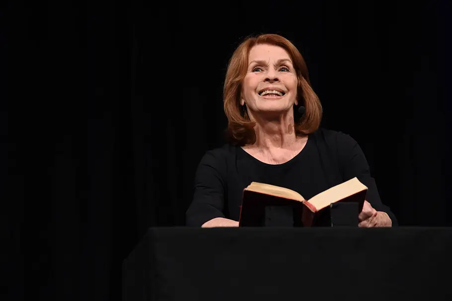 Senta Berger Senta Berger steht vor einem schwarzen Hintergrund und lächelt während die ein Buch auf dem Schoß geöffnet hat.