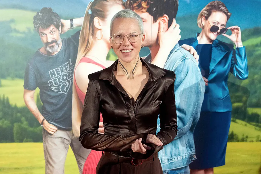 Stefanie Heinzmann steht lächelnd mit kurzen hellen Haaren, Brille und dunkler Satinbluse vor einem Plakat mit mehreren Personen.