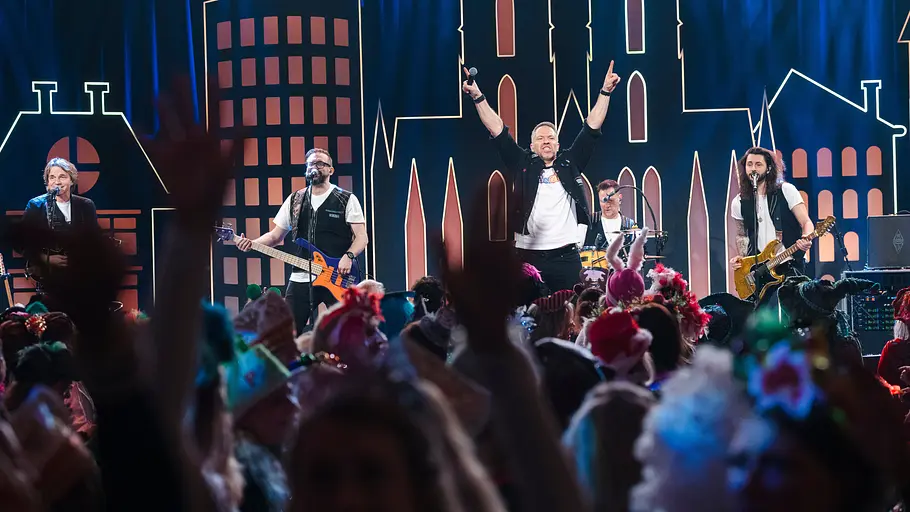 Kölle Alaaf - Die Mädchensitzung: Die sechs Musiker der Musikgruppe Höhner stehen auf der Bühne, der Sänger hat beide Arme nach oben gestreckt und performt.