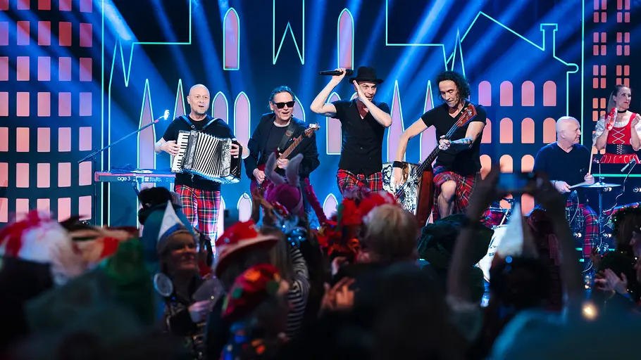 Kölle Alaaf - Die Mädchensitzung: Die fünf Musiker der Musikgruppe Brings stehen auf der Bühne und performen. Vier Musiker halten ihre Instrumente in den Händen, ein Musiker hält als Sänger ein Mikrofon in der Hand.
