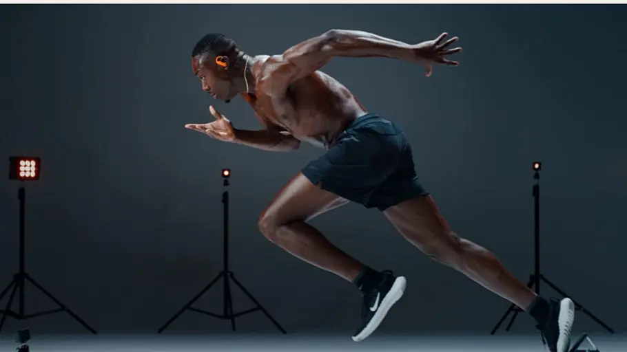 Ein muskulöser Athlet in Sportbekleidung startet mit voller Kraft zu einem Sprint. Im Hintergrund stehen mehrere Scheinwerfer und technische Geräte, die auf eine professionelle Trainingsumgebung hinweisen. Der Läufer trägt kabellose Kopfhörer und fokussiert sich ganz auf seine Bewegung.
