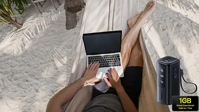 Eine Person arbeitet entspannt mit einem Laptop in einer Hängematte am Sandstrand. Im Vordergrund ist ein mobiler WLAN-Router mit Hinweis auf 1 GB globales Datenvolumen pro Monat zu sehen – Symbol für mobiles Arbeiten und Internetzugang unterwegs. - Foto: istock/anyaberkut/PR