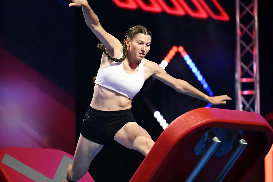 Lana Hohenstein ist Teilnehmer bei Ninja Warrior Germany 2026 Lana Hohenstein balanciert mit ausgestreckten Armen zwischen roten Hindernissen. Sie trägt ein weißes Top und schwarze Shorts.