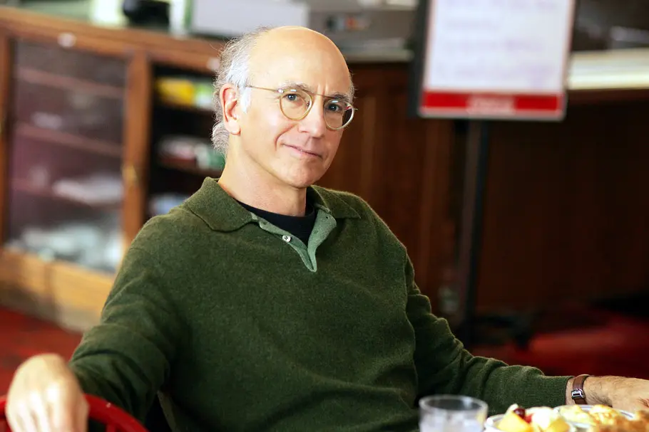 Larry David sitzt auf einem Stuhl und lächelt. Vor ihm steht eine Mahlzeit.