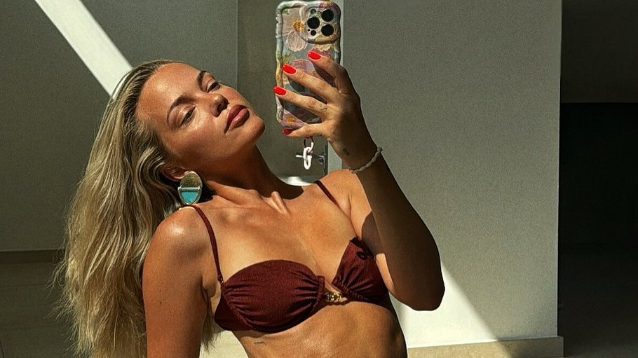 Auf dem Bild sitzt Laura Maria Rypa im Sonnenlicht auf dem Boden und macht ein Selfie mit ihrem Smartphone. Sie trägt einen weinroten Bikini, große runde Ohrringe und lässt ihr blondes Haar über die Schulter fallen. Mit der Pose betont sie ihren trainierten Körper, während das Licht Schatten auf Wand und Boden wirft.