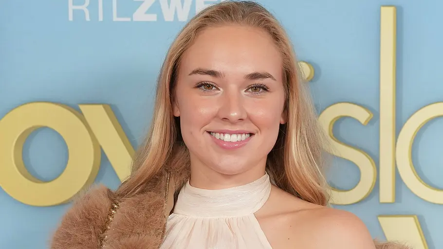Laura Peuker: Eine lächelnde Frau mit langen blonden Haaren trägt ein helles Kleid und eine braune Felljacke vor einer blauen RTLZWEI-Wand.