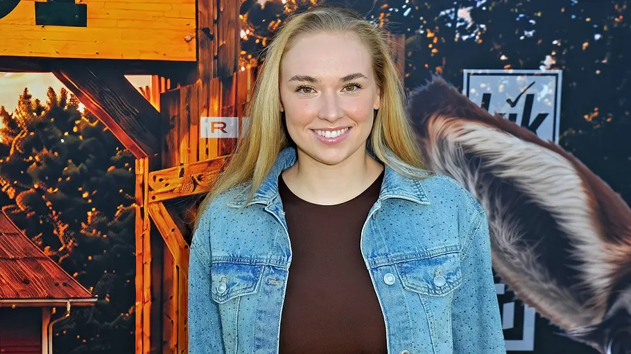 Laura Peuker im Steckbrief. Laura Peuker mit langen blonden Haaren trägt eine blaue Jeansjacke vor einer Kulisse mit Holzhäusern.
