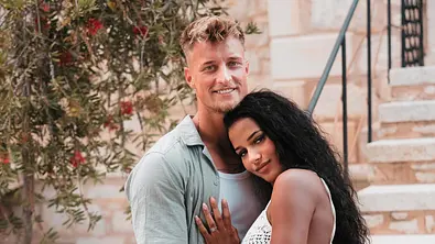 Laurenz und Brenda stehen eng umschlungen vor einer Steintreppe, beide lächeln in die Kamera. Die Frau trägt ein weißes, gehäkeltes Kleid, der Mann ein helles Sommeroutfit. - Foto: RTL