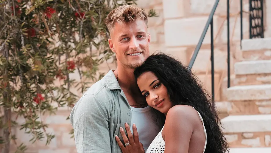 Laurenz und Brenda vor der Teilnahme an Temptation Island VIP Laurenz und Brenda stehen eng umschlungen vor einer Steintreppe, beide lächeln in die Kamera. Die Frau trägt ein weißes, gehäkeltes Kleid, der Mann ein helles Sommeroutfit.