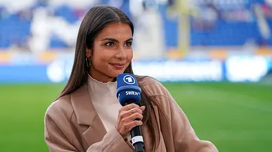 Lea Wagner hält ein blaues SWR-Mikrofon und trägt einen beigen Mantel. Hinter ihr sind Rasen und blaue Stadionränge zu sehen. - Foto: IMAGO / foto2press