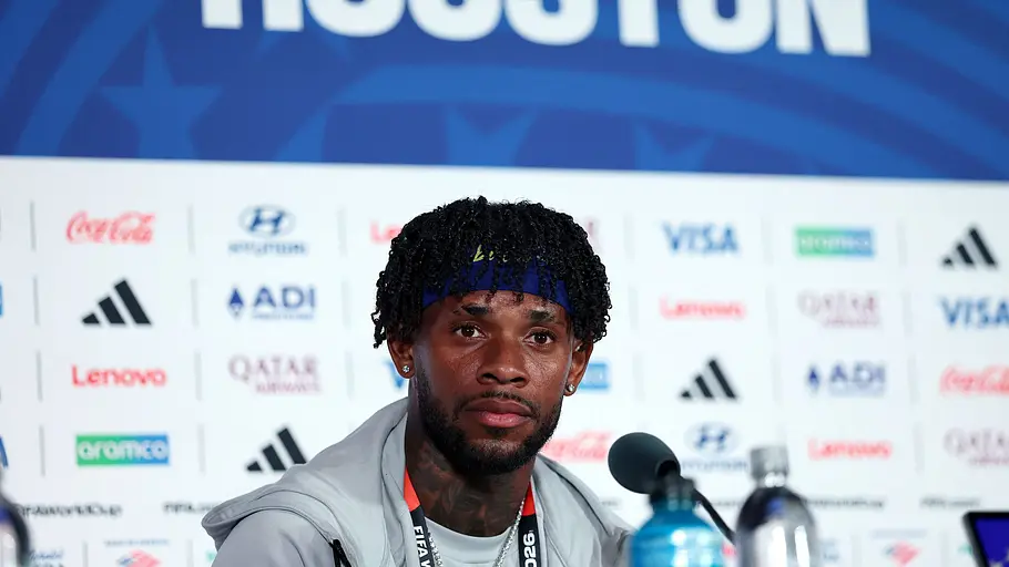Ein Nationalspieler sitzt bei einer Pressekonferenz vor einem Mikrofon. Hinter ihm ist eine Sponsorenwand mit der Aufschrift „Houston“ sowie mehreren Partnerlogos zu sehen.