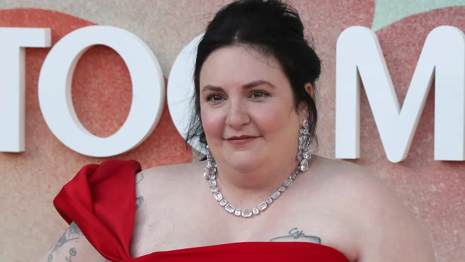 Lena Dunham Lena Dunham trägt ein rotes Kleid mit großer Schulterschleife, eine funkelnde Halskette und lange Ohrringe. Ihre Arme sind tätowiert.