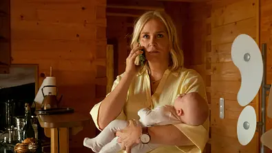 Lena Lorenz – Wahlverwandtschaft: Lena Lorenz (Judith Hoersch) hält ein Baby im Arm und telefoniert mit besorgtem Gesichtsausdruck mit einem Handy. Sie befindet sich in einer Küche, deren Wände mit Holz vertäfelt sind. - Foto: ZDF und Erika Hauri